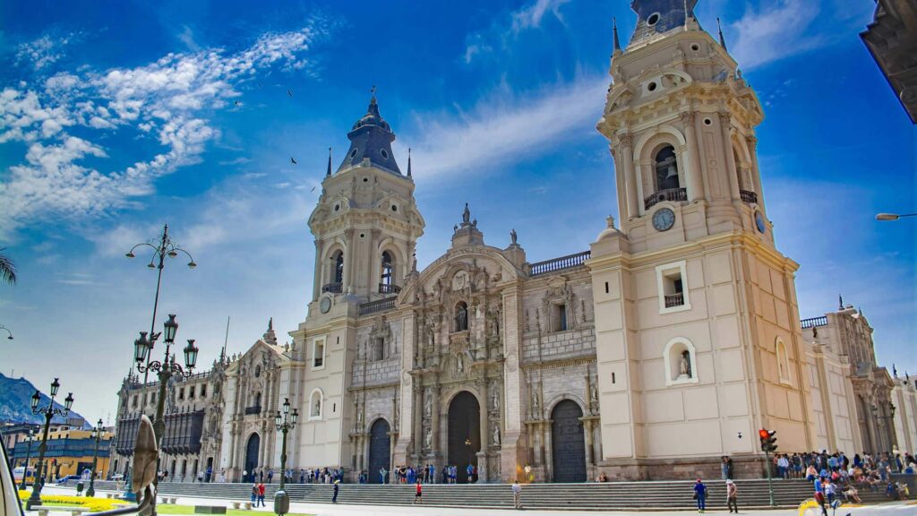 The Lima Cathedral's ornate facade, twin towers, and blue-domed cupola rise against a bright blue sky with scattered clouds.