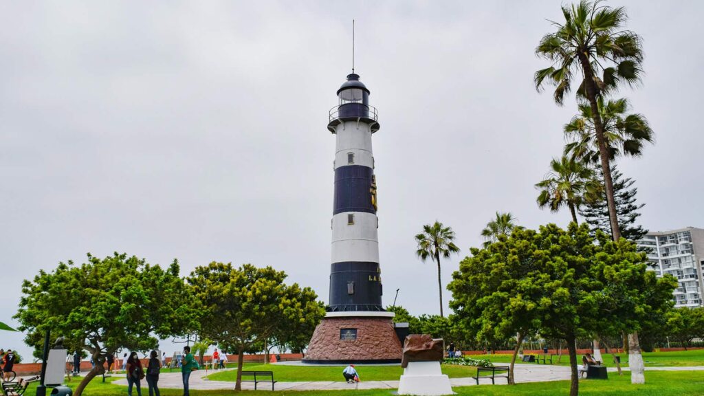 Striking black and white striped lighthouse stands tall in a lush green park with palm trees against a cloudy sky.