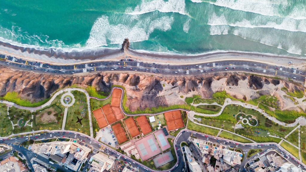 Aerial view of Lima, Peru's coastline featuring turquoise ocean waves, a busy highway, dramatic cliffs, lush green parks, tennis courts.
