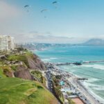 Scenic aerial view of Miraflores, Lima, Peru, featuring paragliders soaring over the coastline, vibrant green cliffs.