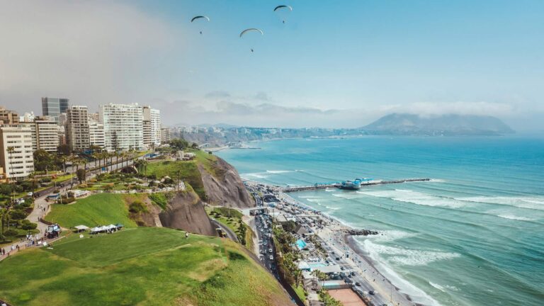 Scenic aerial view of Miraflores, Lima, Peru, featuring paragliders soaring over the coastline, vibrant green cliffs.